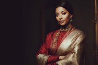 Young South Asian woman in traditional red and gold silk saree with ornate jewelry posing elegantly against dark background for cultural fashion portrait.