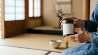 Senior man with brown skin tone holding white takeout coffee cup in modern home interior with wooden doors and natural light.