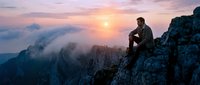 Man sitting on rocky mountain cliff at sunrise overlooking misty valley landscape. Outdoor adventure and nature exploration concept for travel, hiking, and lifestyle design projects.