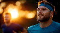Athletic bearded man in blue headband and shirt running outdoors during golden sunset with blurred jogger in background, warm evening workout atmosphere.