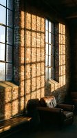 Sunlit industrial loft interior with vintage leather armchair positioned near large factory windows casting geometric shadows on exposed brick walls, creating warm golden light ambiance for modern urban living spaces.