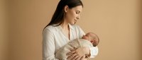 Young Asian mother in white shirt tenderly holding and breastfeeding her newborn baby in warm beige studio setting for maternal bonding and childcare concepts.