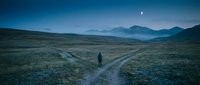 Lone traveler walking on remote mountain road at twilight with crescent moon overhead. Atmospheric landscape perfect for adventure and solitude concepts.