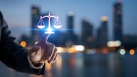 Businessman hand holding glowing justice scales icon against blurred city skyline at dusk. Legal services, law firm, attorney consultation concept.