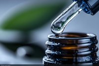 Clear liquid dropping from glass dropper into dark bottle, macro photography with selective focus showing essential oil or medicine against blurred green background.
