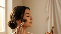 Young woman applying makeup with brush in soft natural light by window. Morning beauty routine skincare cosmetics preparation for special occasion.