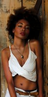 Young African American woman with natural afro hair wearing white crop top and pendant necklace poses against rustic wooden background, expressing confidence and style.