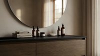 Minimalist bathroom vanity with dark wood cabinet, round mirror, and amber glass bottles arranged on countertop near window with neutral curtains.