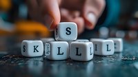 White letter cubes spelling out skills word on textured blue surface with blurred background and hand placing cube, representing personal development and learning concept.