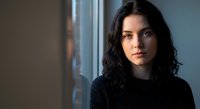 Young brunette woman with wavy hair wearing black sweater looking thoughtfully at camera near window with soft natural lighting for portrait photography.