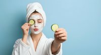 Young woman in white bathrobe and towel applies facial mask while holding cucumber slices for natural skincare routine and spa treatment at home.