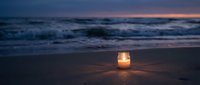 Glowing candle lantern on sandy beach at twilight with ocean waves and purple sunset sky creating romantic peaceful atmosphere for meditation relaxation.