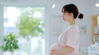 Young Asian pregnant woman in pink dress standing near window, gently holding belly with peaceful expression in bright modern interior with plants.