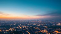 Aerial panoramic view of illuminated cityscape at dusk with glowing streets and buildings under dramatic sunset sky, creating atmospheric urban landscape.