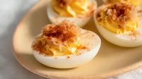 Creamy deviled eggs garnished with crispy bacon bits and paprika powder served on ceramic plate, close up view of classic appetizer with smooth filling.
