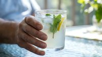 Closeup of male hand holding glass of refreshing lemonade with fresh mint and lemon slices on sunny outdoor patio, perfect for beverage, summer drink, and lifestyle content.
