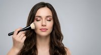 Young brunette woman applying makeup with brush on face against gray background for beauty and cosmetics routine.