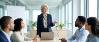 Mature woman with gray hair, Caucasian ethnicity, wearing business attire, leading a corporate meeting with diverse colleagues in a modern office environment with natural light and plants.