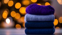 Stack of cozy knitted winter sweaters in blue and purple shades on blurred golden bokeh background, seasonal fashion and warmth concept.