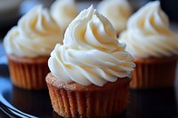 Freshly baked vanilla cupcakes topped with swirled cream cheese frosting on dark plate, macro food photography showing texture and details of homemade dessert.