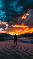 Silhouetted person standing on sand dunes at dramatic sunset with fiery orange sky and turquoise storm clouds over mountain landscape. Contemplative solitary moment.