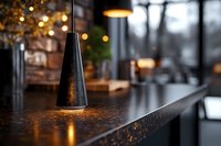 Modern black cone pendant lamp illuminating dark granite countertop in contemporary restaurant interior with soft bokeh lights and moody atmosphere.