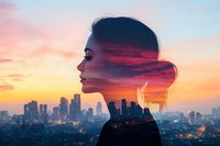 Double exposure portrait of young Asian woman silhouette combined with vibrant sunset cityscape showing illuminated urban skyline and dramatic orange sky.