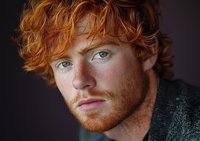 Young man with bright ginger curly hair and freckles wearing green sweater gazes intensely at camera in dramatic moody portrait against dark background.