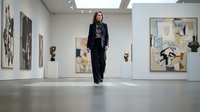 Woman in black suit walking through contemporary art gallery with modern paintings and sculptures on display.