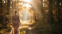 Woman in floral dress standing on forest path during golden hour sunlight, surrounded by autumn trees and natural woodland scenery for editorial and lifestyle use.