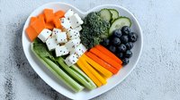 Fresh vegetables and berries arranged in heart shaped plate with feta cheese cubes, colorful bell peppers, celery, carrots, cucumber, broccoli and blueberries on gray background.