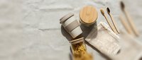 Flat lay composition featuring stainless steel containers, wooden lid, spoons, and linen cloth on neutral textured surface for lifestyle and product photography.