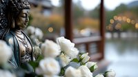 Serene Buddha statue with white roses in foreground, soft bokeh lights creating peaceful zen atmosphere. Meditation and mindfulness concept for spa and wellness.