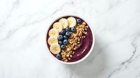 Purple acai smoothie bowl topped with fresh banana slices, blueberries and granola on marble background for healthy breakfast and nutrition content.