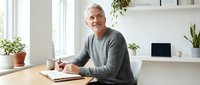 Mature man with gray hair and light complexion, wearing casual gray sweater, writing notes at wooden desk in bright modern office with laptop and plants.