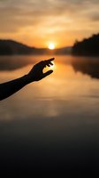 Silhouette of hand reaching toward golden sunset over calm lake water with warm orange sky and mountain reflections for spiritual wellness concepts.