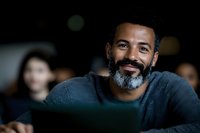 Smiling mature Black man with salt and pepper beard wearing blue sweater in dark room with computer screen glow, representing remote work or education setting.
