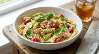 Fresh pasta salad with rotini, cherry tomatoes, basil and herbs in white bowl on wooden table with iced tea glass for summer dining and meal preparation.