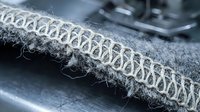 Industrial sewing machine creating decorative overlock stitch on gray fabric edge. Close-up macro view of textile manufacturing process in garment production.