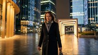 Professional businesswoman walking through modern urban plaza at night with illuminated skyscrapers and corporate buildings creating dynamic cityscape backdrop.