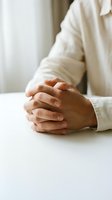 Clasped hands of a person resting on a white table, conveying focus, patience, or prayer in a calm, minimal setting.