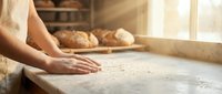 Baker kneading dough on wooden surface with fresh bread loaves in background, artisan bakery production and food preparation.