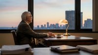 Senior businessman with glasses and gray hair, wearing dark business suit, sitting at wooden desk in high-rise office during sunset. Cityscape visible through floor-to-ceiling windows with golden hour lighting. Desk lamp illuminates workspace with documents and office supplies. Professional corporate environment for business, leadership, success themes.