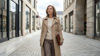 Confident mature businesswoman in beige trench coat walking through modern urban shopping district. Professional female executive in elegant outerwear on city street.
