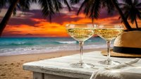 Two champagne glasses on tropical beach table at sunset with palm trees and ocean waves creating romantic vacation atmosphere for celebration.