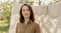 Young brunette woman in brown shirt standing outdoors near clothesline with natural linen fabrics drying in spring garden with blooming tree branches.
