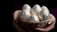 Decorative white Easter eggs with golden geometric pattern held in woven basket. Dark moody background creates elegant holiday composition with feminine hands.