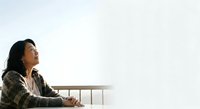 Young Asian woman with long dark hair wearing a patterned cardigan, leaning on a wooden railing against a white sky background, contemplative expression, suitable for lifestyle and wellness content.