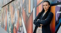 Young woman with dark hair and olive complexion wearing black blazer and beige pants standing confidently with crossed arms against colorful urban street art mural backdrop.