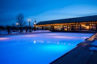 Illuminated outdoor swimming pool at dusk with glowing blue water reflections and warm lights from modern resort building, creating serene winter atmosphere.